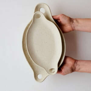 Pair of oatmeal -colored porcelain offering platters with sculptural cut-outs and speckled glaze, displayed stacked.