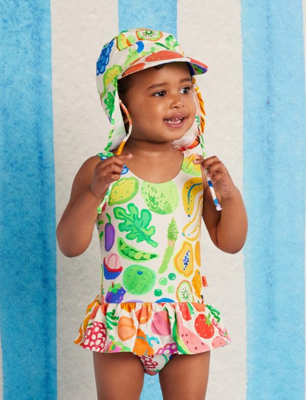 Model wears Halcyon Nights Baby Frill Bather in Eat The Rainbow print—frill-trimmed swimsuit with vibrant rainbow artwork and built-in nappy liner for babies