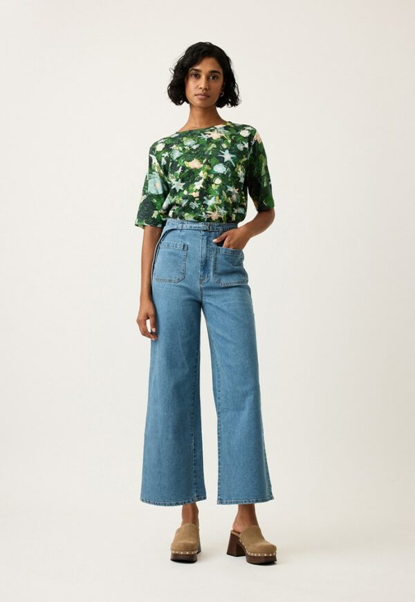 Model wearing Nancybird Briar Pants in denim—high‑waist straight‑leg jeans with self‑fabric tie belt, front and back patch pockets, and contrast top stitch detailing in mid wash denim