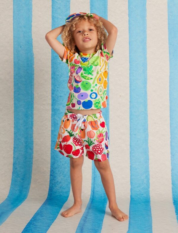 Model wears Halcyon Nights Kids Boardshorts in Eat The Rainbow print—colourful quick-dry shorts with elastic waist, drawcord, and Seeun Park’s vibrant artwork